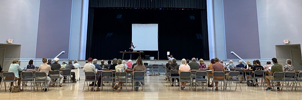 Picture of a poll worker class being taught in a large room with a stage. A woman stands on the stage speaking to an audience of about 35 people..